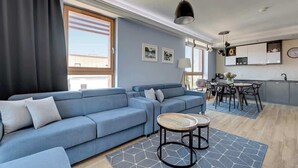 A modern living room with a blue sofa, a dining area with a table and chairs, and a kitchen with cabinets and a clock. - Aura 222 | River View (Gdańsk)
