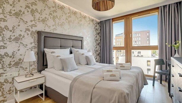 A modern hotel room with a large bed, a bedside table, a dresser, and a view of the city. - Aura 222 | River View (Gdańsk)