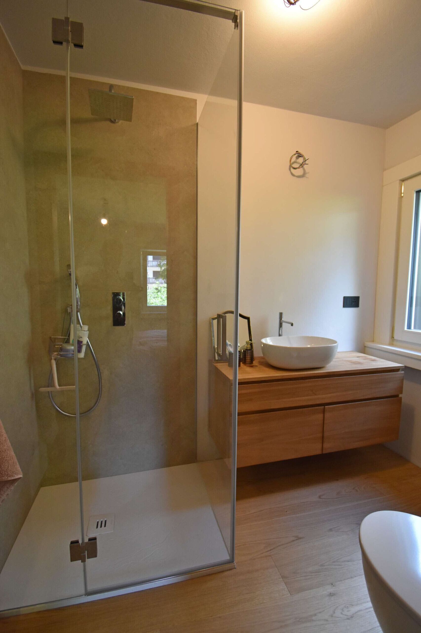 Ein modernes Badezimmer mit einer begehbaren Glasdusche, einem Holzwaschtisch mit weißem Waschbecken und einem Fenster für natürliches Licht.