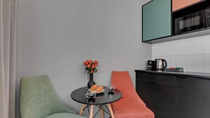 Ett litet pentry med en svart bänkskiva, en kaffebryggare och en mikrovågsugn. Där finns ett runt bord med två stolar, en grön och en röd, och en vas med röda blommor på bordet.