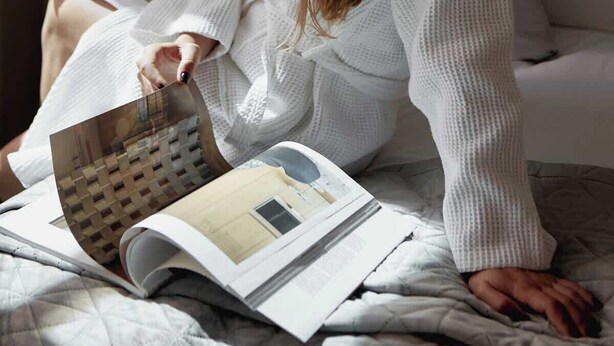 Een persoon zit op een bed met een tijdschrift, in een kamer met een bed, kussens en een raam.