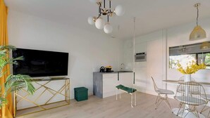 Una sala moderna con televisor de pantalla plana, un comedor con mesa y sillas redondas, una cocina con lámpara colgante y un amplio ventanal con flores amarillas.
