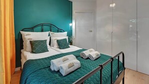 Habitación de hotel con una pared de acento color aguamarina, una cama con ropa blanca y almohadas verdes, estructura de cama metálica, un armario y una puerta.