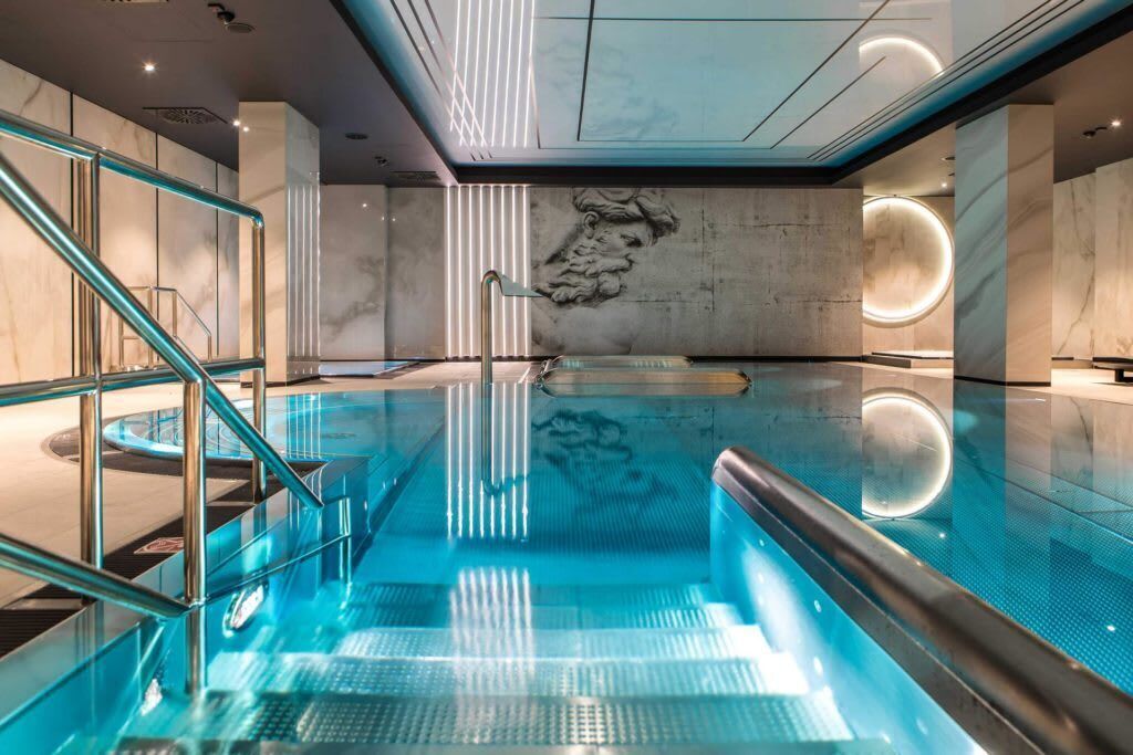 Ein moderner Innenpool mit Glastrennwand, einer Marmorwand mit Reliefbild und einer runden Leuchte.
