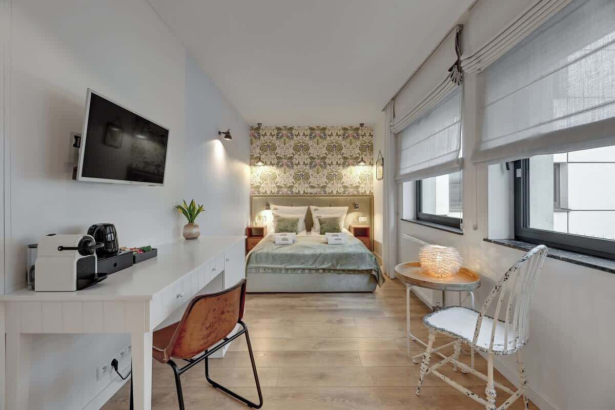 Ein modernes Schlafzimmer mit einem Bett, Nachttischen, einem Fernseher, einem Schreibtisch mit einer Kaffeemaschine, einem Sessel und einem Fenster mit Jalousien.