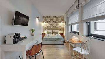 Ein modernes Schlafzimmer mit einem Bett, Nachttischen, einem Fernseher, einem Schreibtisch mit einer Kaffeemaschine, einem Sessel und einem Fenster mit Jalousien.