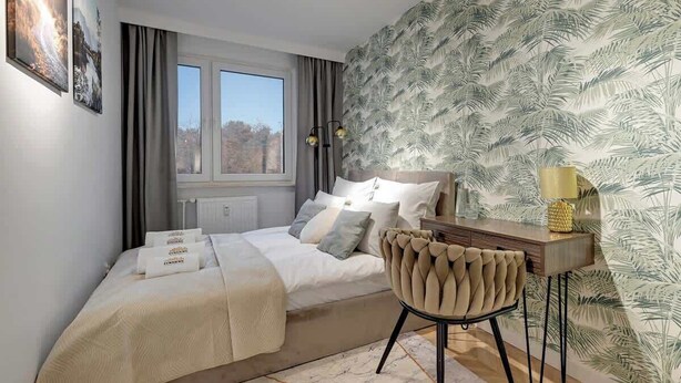 Ein Schlafzimmer mit einem Bett, einem Schreibtisch, einem Stuhl, einem Fenster und einer Wand mit Blätermuster.