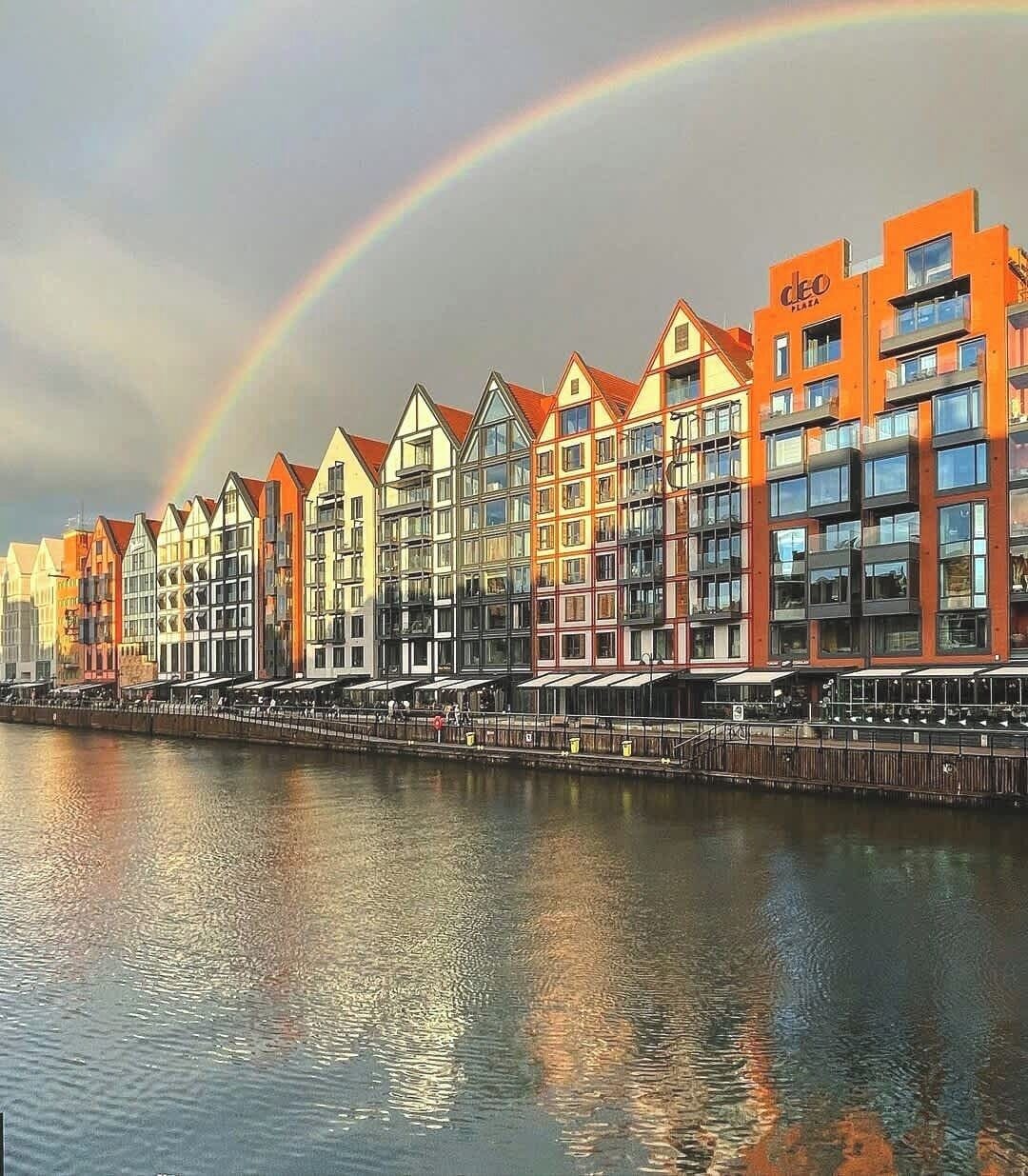 Eine Reihe bunter Gebäude entlang eines Ufers, am Himmel ein Regenbogen.
