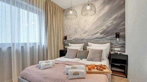 Une chambre d’hôtel moderne avec un grand lit, deux tables de chevet, une petite table d’appoint, une fresque murale et une fenêtre agrémentée de rideaux légers.