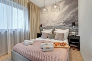 Ein modernes Hotelzimmer mit einem großen Bett, zwei Nachttischen, einer Ablagefläche, einem Wandbild und einem Fenster mit durchsichtigen Vorhängen.