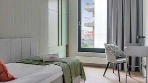 Et moderne hotelværelse med seng, skrivebord, stol og stort vindue med udsigt til bygninger.
