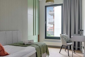 A modern hotel room with a bed, a desk, a chair, and a large window with a view of buildings. - Brabank 10 | Riverside & Balcony | Parking (Gdańsk)