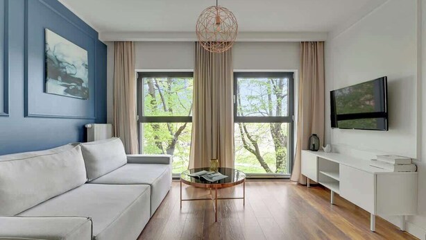 A modern living room with a white sofa, a glass coffee table, a TV on the wall, and large windows with curtains. - Rezydencja Wintera 11 | Riverside (Gdańsk)