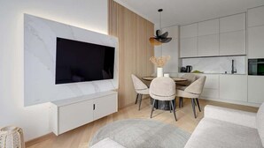 Un soggiorno moderno con una televisione a schermo piatto, un'area pranzo con un tavolo rotondo e sedie, e una cucina con mobili bianchi.