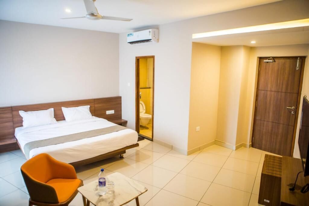 Una habitación de hotel con una cama, un sillón naranja, una mesita con una botella, un televisor y un equipo de aire acondicionado.