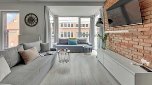 Een moderne woonkamer met een grijze bank, een salontafel, een televisie bevestigd aan een bakstenen muur, en een groot raam met uitzicht op gebouwen.
