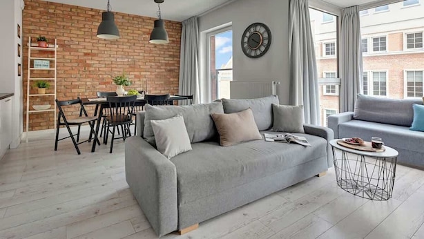 Een moderne woonkamer met een grijze bank, een eettafel met stoelen, een bijzettafeltje en een metselwerk muur.