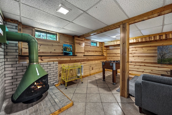 A cozy room with a fireplace, a foosball table, and a window with a view of trees.