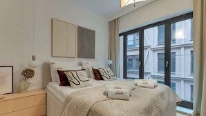 Une chambre d’hôtel moderne avec un grand lit, une table de chevet, une lampe fixée au mur et une grande fenêtre donnant sur la ville.