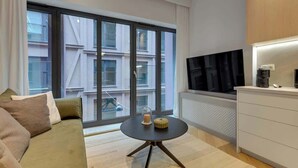 Un salon moderne comprenant un canapé, une table ronde avec une bougie et un objet décoratif, un téléviseur et un coin cuisine avec un four à micro-ondes.