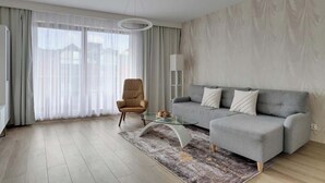 Ein modernes Wohnzimmer mit grauem Sofa, einem Glastisch, einem beigefarbenen Sessel und transparenten Vorhängen.