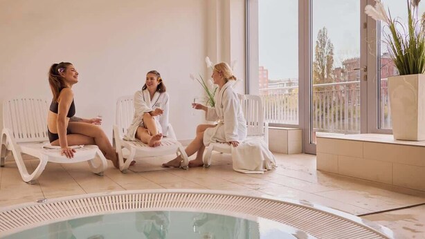 Drei Personen sitzen an Liegestühlen an einem Pool in einem modernen Innenbereich mit großen Fenstern.