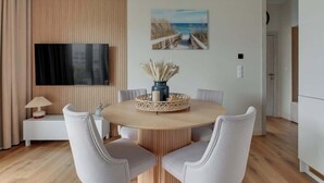 Et moderne spiseområde med et rundt bord, hvide stole, et tv ophængt på væggen og et maleri med strandmotiv.