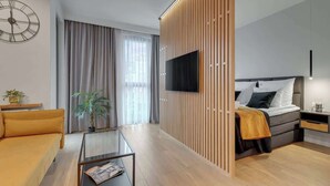 Moderni hotellihuone, jossa on sohva, sohvapöytä, televisio, sängynpääty ja puinen sermi.