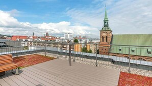 Kattoterassi, jossa on puinen penkki, metallinen kaide ja näkymä kaupunkimaisemaan, johon kuuluu myös vihreä torni omaava kirkko.