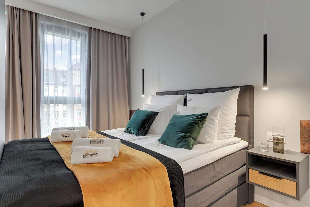 A hotel room with a bed, a nightstand, a window with curtains, and a wall-mounted light fixture.