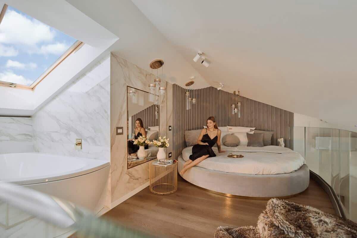 A modern bedroom with a skylight, a round bed, a large mirror, and a bathroom with a white bathtub.