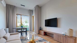 Ein modernes Wohnzimmer mit einer Couch, einem Glastisch, einem Flachbildfernseher, einem Essbereich mit Tisch und Stühlen sowie einem großen Fenster mit Vorhängen.