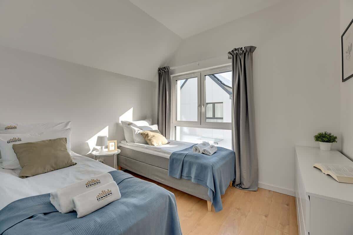 A bedroom with two beds, a window with curtains, a bedside table with a lamp, and a small plant.