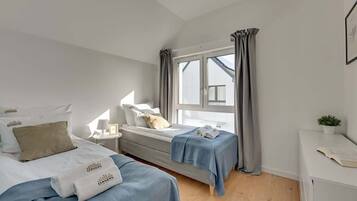 A bedroom with two beds, a window with curtains, a bedside table with a lamp, and a small plant.