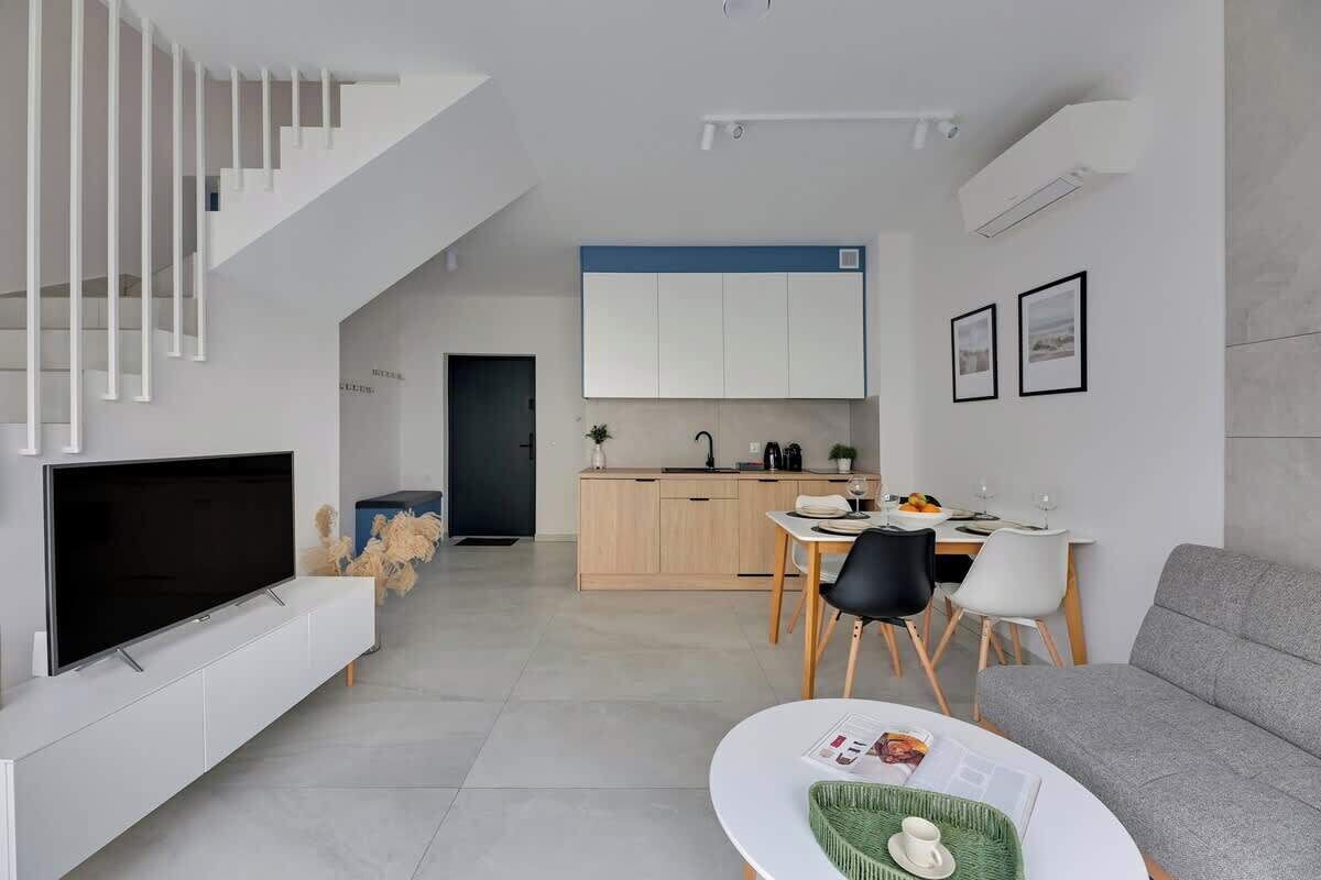 Ein modernes Wohnzimmer mit Flachbildfernseher, grauem Sofa, Essbereich mit Tisch und Stühlen sowie einer Küche mit Holzausstattung und blauer Akzentwand.