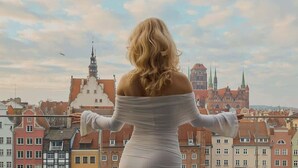 Une femme vêtue d’une robe blanche se tient sur un balcon d’où l’on aperçoit une vue panoramique sur la ville, avec ses bâtiments historiques.