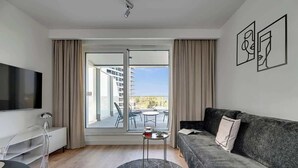 Ein modernes Wohnzimmer mit einem Flachbildfernseher, einem grauen Sofa, einem runden Couchtisch und Blick auf einen Balkon mit Gartenmöbeln.