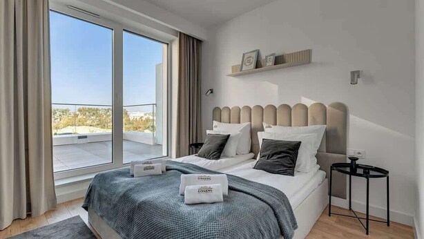 Ein modernes Hotelzimmer mit einem großen Bett, einem kleinen Nachttisch, einem Fenster mit Vorhängen und Blick ins Freie.