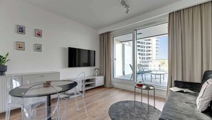 Ein modernes Wohnzimmer mit einem Glastisch, einer Couch, einem Fernseher und einem Balkon mit Blick auf Gebäude.