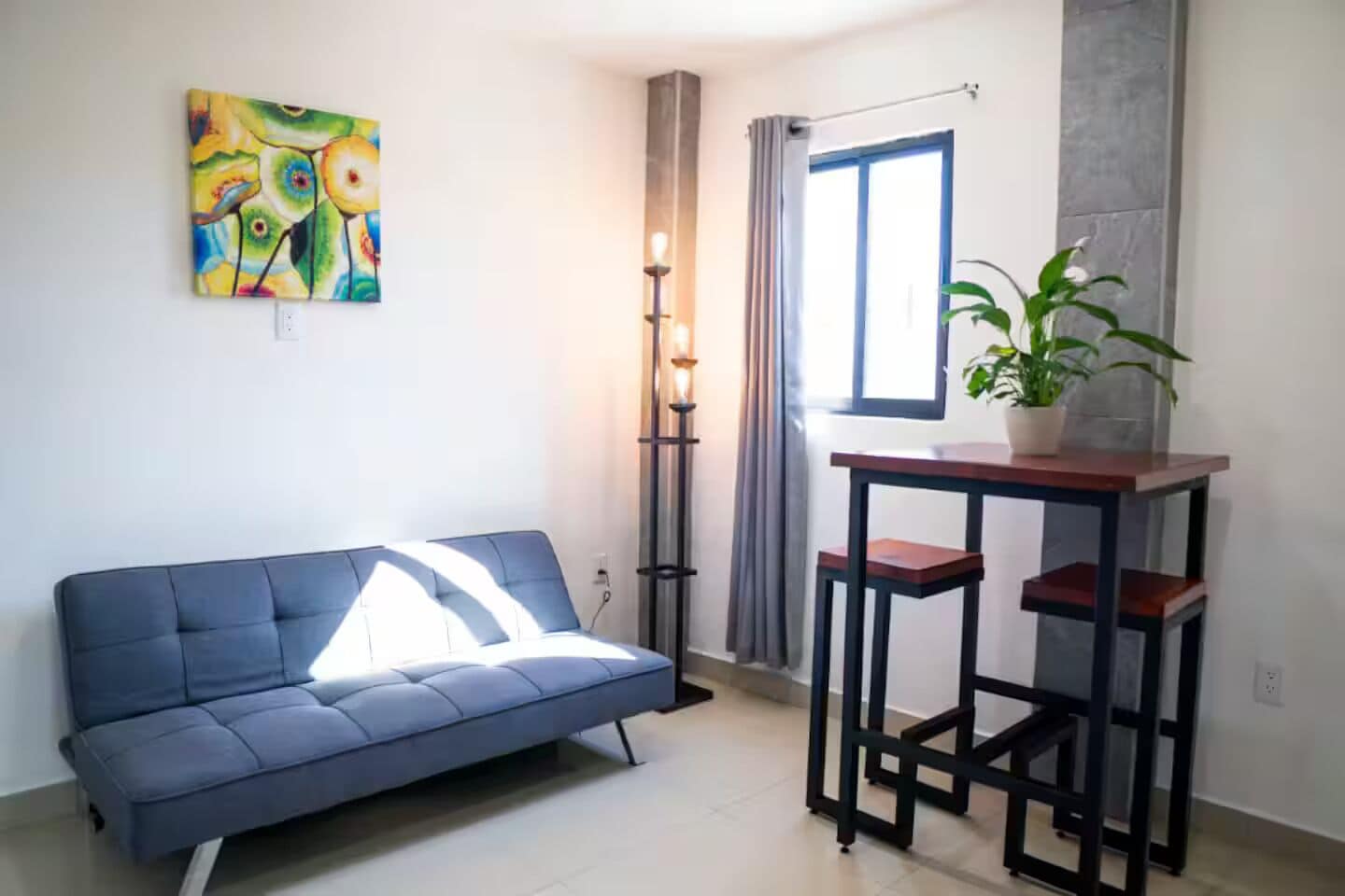 Ein Zimmer mit einem blauen Sofa, einem kleinen Tisch mit zwei Hocker, einer Stehlampe, einem Blumentopf und einem Fenster mit Vorhängen.