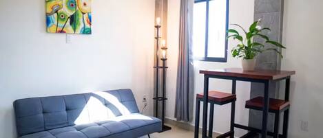 Una habitación con un sofá azul, una pequeña mesa con dos taburetes, una lámpara de pie, una planta en maceta y una ventana con cortinas.