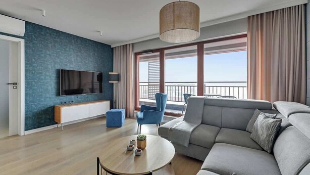 A modern living room with a large flat-screen TV, a sofa, a coffee table, and a view of the city through large windows. - Nº 83 with Balcony | Pool, SPA & Gym | River View (Gdańsk)