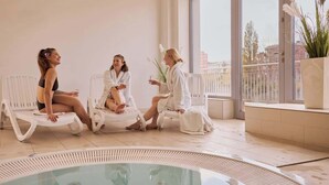 Three individuals seated on lounge chairs by a poolside in a modern indoor setting with large windows. - Nº 83 with Balcony | Pool, SPA & Gym | River View (Gdańsk)