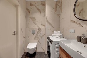 A modern bathroom with a marble wall, a washing machine, a toilet, and a sink with a mirror. - Scala 37 with Balcony | City Center | Parking (Gdańsk)