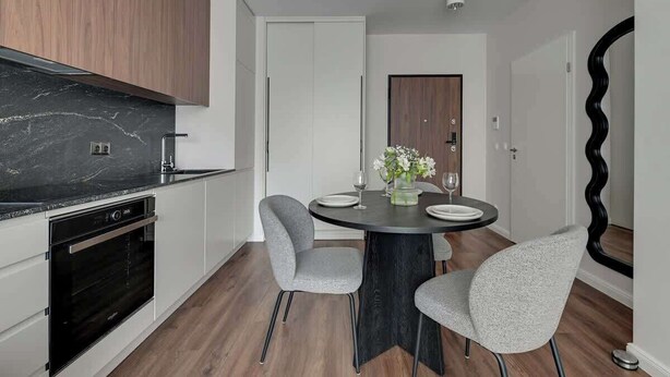 A modern kitchen with a black marble countertop, white cabinets, and a built-in oven. There is a dining area with a round table, four grey chairs, and a vase with flowers. - Scala 37 with Balcony | City Center | Parking (Gdańsk)