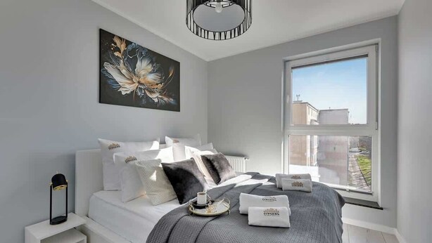Ein modernes Schlafzimmer mit großem Fenster, einem Bett mit vielen Kissen, einem Nachttisch mit Lampe und einem Bild an der Wand.