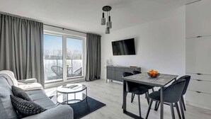 Ein modernes Wohnzimmer mit einer Couch, einem Esstisch, Stühlen, einem an der Wand montierten Fernseher und einem großen Fenster mit Vorhängen.