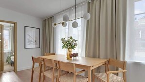 Uma área de refeições com mesa e cadeiras de madeira, um vaso com flores e uma janela com cortinas.