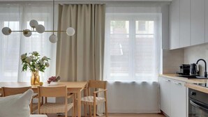 Uma cozinha moderna com armários brancos, uma área de refeições com mesa e cadeiras de madeira, e uma janela grande com cortinas.