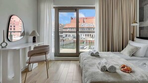 A hotel room with a bed, a desk, a chair, a window with a view of buildings, and a lamp. - Waterlane E2 | Spa, Pool & Gym | Riverside (Gdańsk)